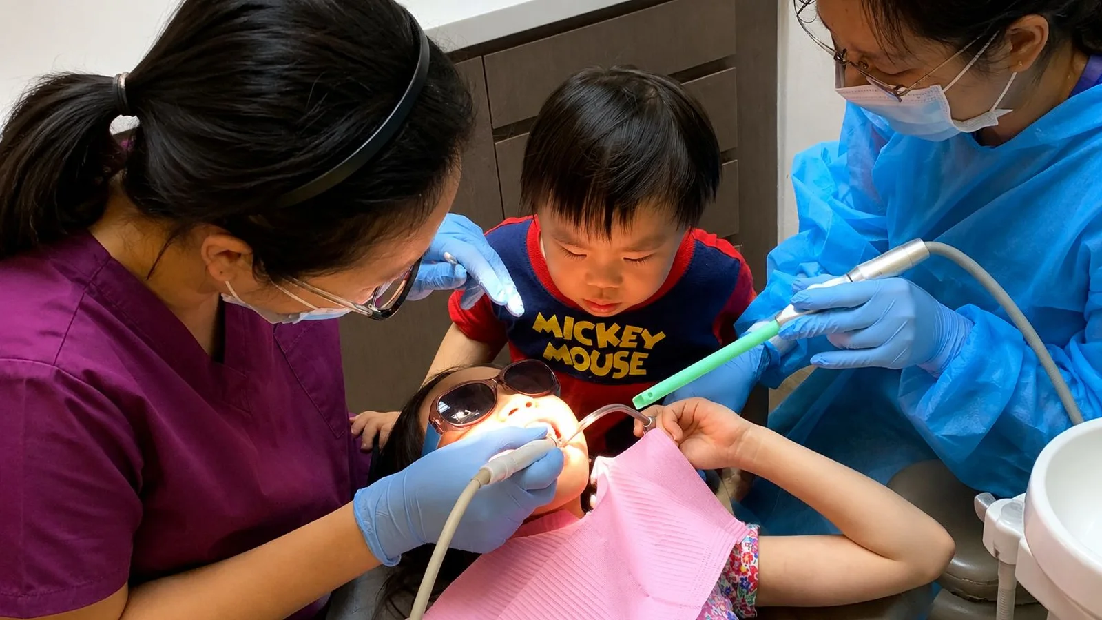 A young child watching curiously as Caring Dental's team gently treats his sister in the dental chair